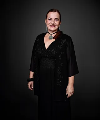 Angelica Leutwiler é uma mulher branca, com cabelo longo e ruivo. Ela está usando um vestido preto e acessórios prateados.