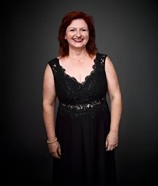 Mônica Weber é uma mulher branca, com cabelo ruivo e longo. Ela está usando um vestido preto e um colar prateado com pingente de gato.