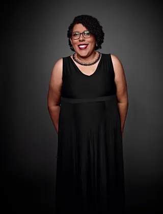 Solange Ferreira é uma mulher negra, com cabelo curto e cacheado. Ela está usando um vestido preto, um colar de contas e óculos.