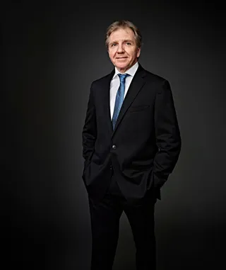 Thierry Fischer é um homem branco de cabelos grisalhos. Ele veste camisa branca, paletó preto e gravata azul.