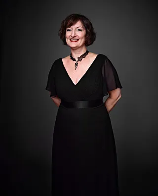 Vesna é uma mulher branca, com cabelo curto e castanho e olhos azuis. Ela está usando um vestido preto e um colar e brincos com detalhes prateados.