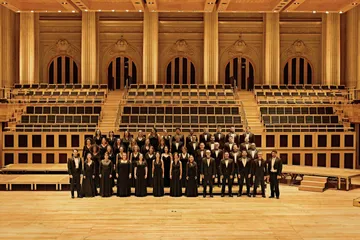 Os músicos do Coro estão posicionados no palco, todos vestem preto e sorriem. 