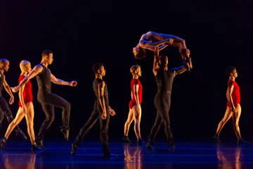 Bailarinos de roupas pretas e bailarinas de maiô vermelho caminham no palco com fundo escuro. No centro, uma bailarina está levantada pelos braços e pernas por um bailarino.
