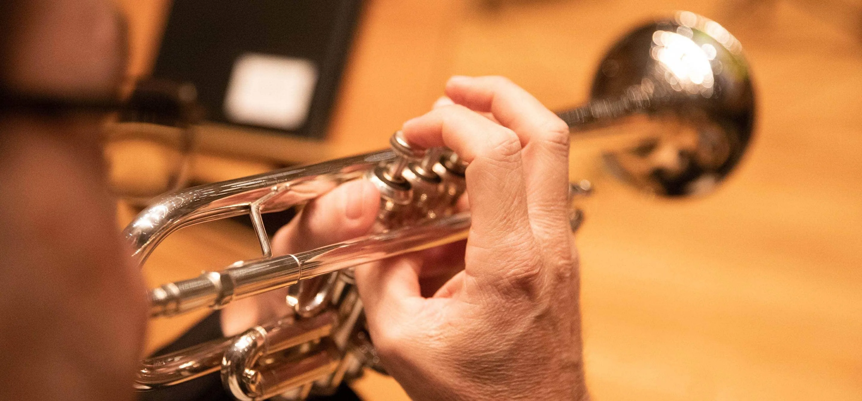 A imagem mostra um trompete dourado, instrumento de metal, sendo tocado por um músico.. 